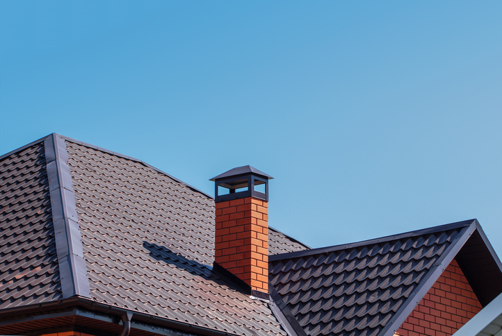 Brick chimney pipe on metal roof of a private house against the sky A modern rooftop with dark gray metal tiles and a red brick chimney topped with a black cap. The clean lines and bright blue sky create a sleek and minimalistic architectural appearance.